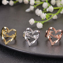 Trio of gold, silver, and rose gold heart name rings displayed on dark plate next to white flowers.
