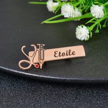 Rose gold custom nurse brooch featuring stethoscope charm with red crystal accent, syringe charm, and engraved name Etoile on rectangular plate.